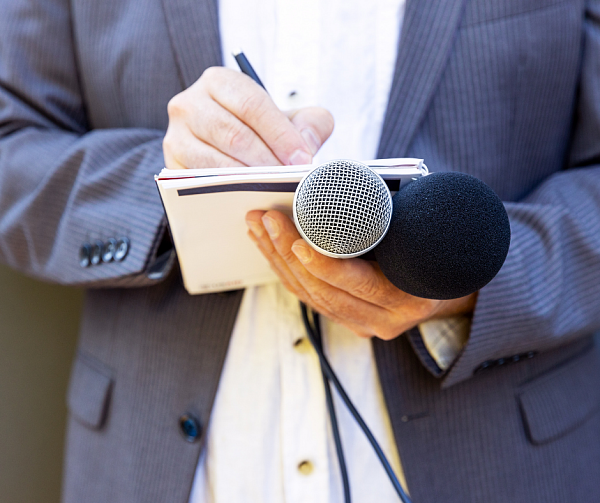 Symbolbild - Ein Journalist schreibt etwas in einem Block und hält zusätzlich zwei Mikrofone in der Hand